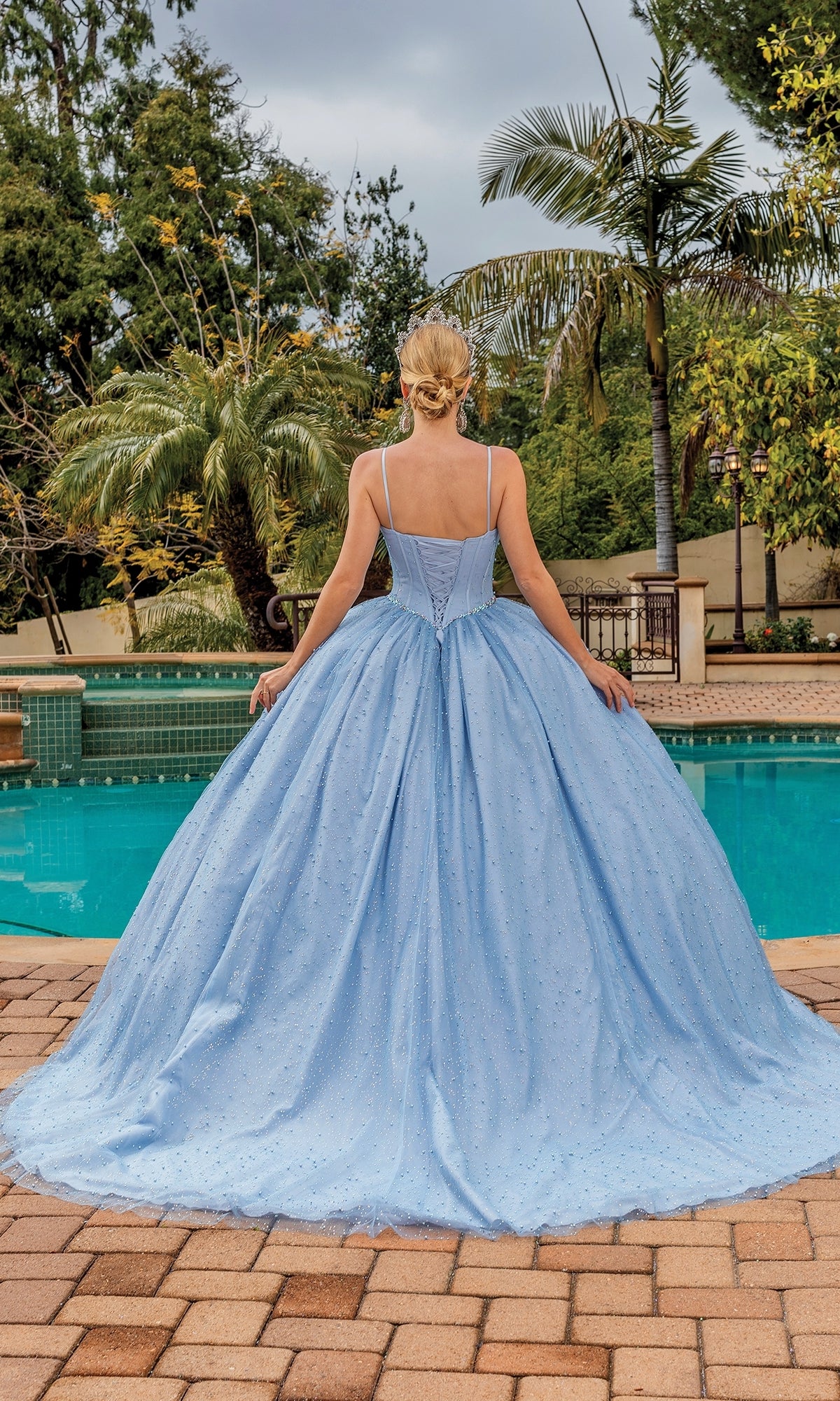 A woman wearing the Quinceanera Dress 1891 By Dancing Queen, featuring a pastel ball gown with a full skirt and sweetheart neckline, stands by a pool with trees and palm plants creating a peaceful backdrop.
